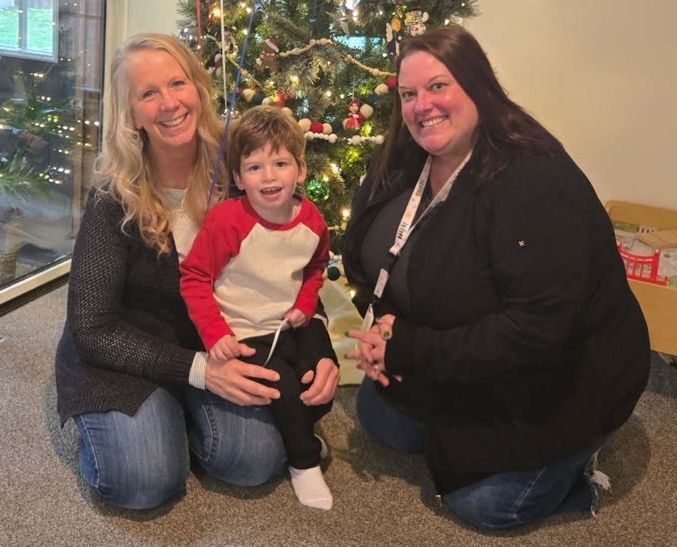 A young child, a family member, and a staff person posing in front of a Christmas tree indoors