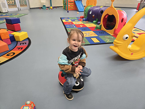 Little girl sitting on a toy that resembles a ladybug in a playroom full of colorful toys